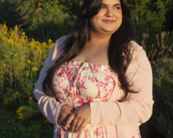 a girl standing near a bushy area near a pond, looking toawards her right side smiling and crossing hands, golden light is hitting her face.